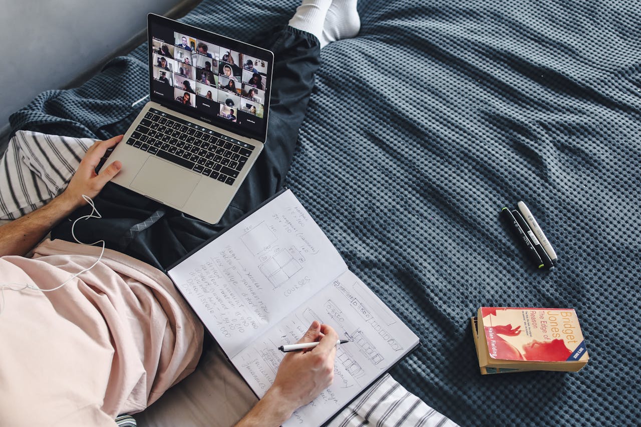 Adult engaged in online class with a laptop and notebook while sitting in a cozy bed setting.